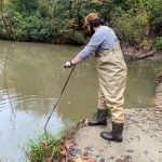 Steve stands on a creek bank wearing waders and boots. He uses a long measurement tool to take a sample of the creek bed