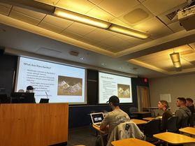 View from the back of a geology classroom looking toward two presentation screens. Shikha Sharma stands behind a podium delivering the lecture.